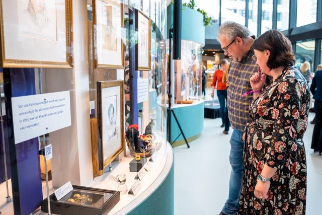 Een man en een vrouw rechts in beeld staan voor een staande vitrine, waarin documenten en portretten zichtbaar zijn.