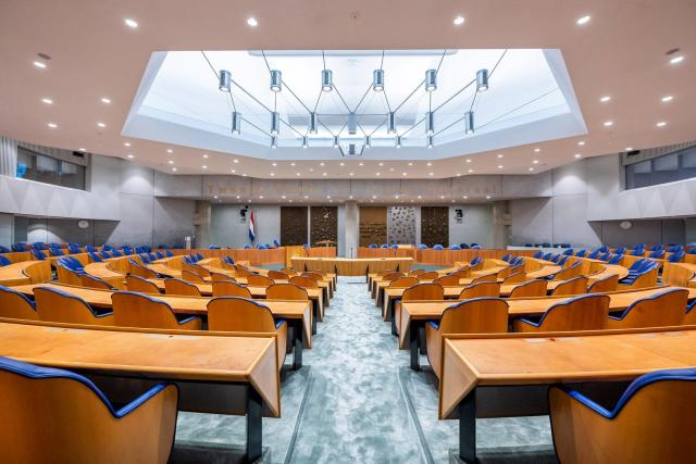 Plenaire zaal leeg, vanachter de Kamerzetels gezien. In het plafond is een groot lichtarmatuur te zien.