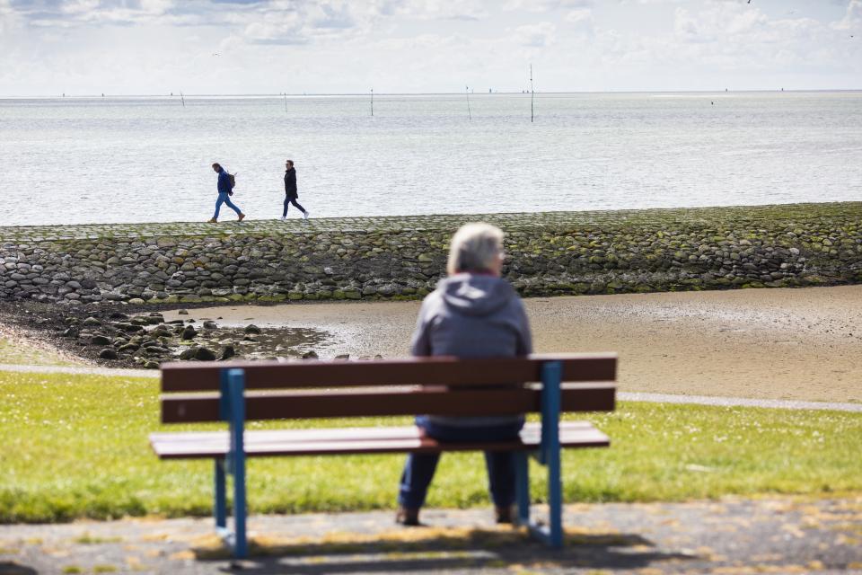 Oudere op bankje met uitzicht op water, van de achterkant gefotografeerd. 