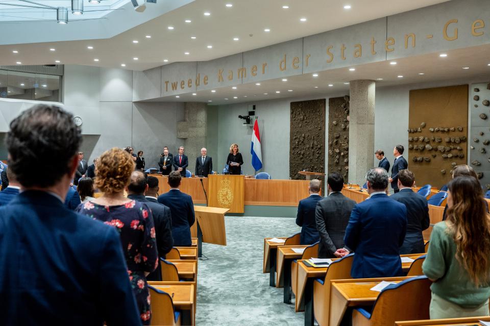 De Kamerleden, Kamervoorzitter, waarnemend Griffier en kabinetsleden staan en houden een moment stilte voor de overleden journalist.