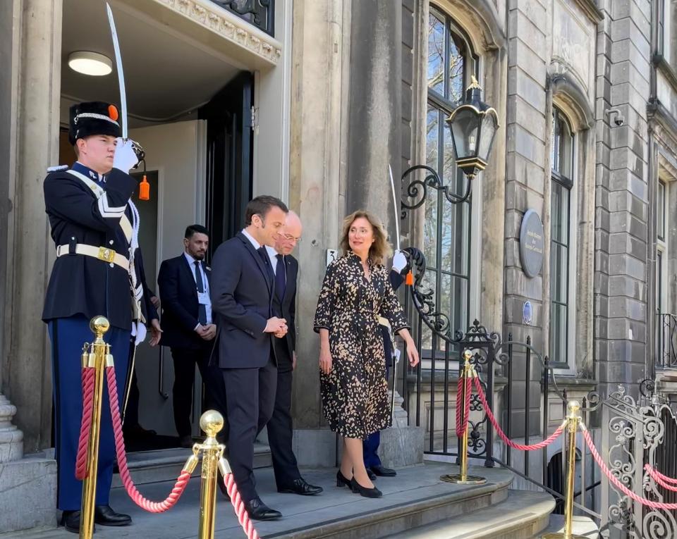 Kamervoorzitter Vera Bergkamp loopt naast Emmanuel Macron het bordes af van de tijdelijke huisvesting van de Eerste kamer. Naast hen heft een man in uniform een sabel. 