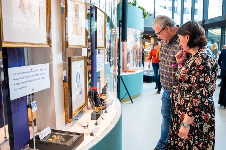 Een man en een vrouw rechts in beeld staan voor een staande vitrine, waarin documenten en portretten zichtbaar zijn. 