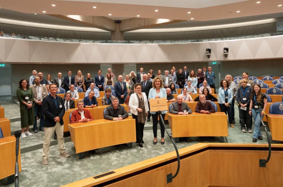 Tussen de bankjes in de plenaire vergaderzaal van de Tweede Kamer houdt Kamervoorzitter Vera Bergkamp het koffertje met opschrift Verantwoordingsdag omhoog, enkele tientallen journalisten staan en zitten om haar heen.