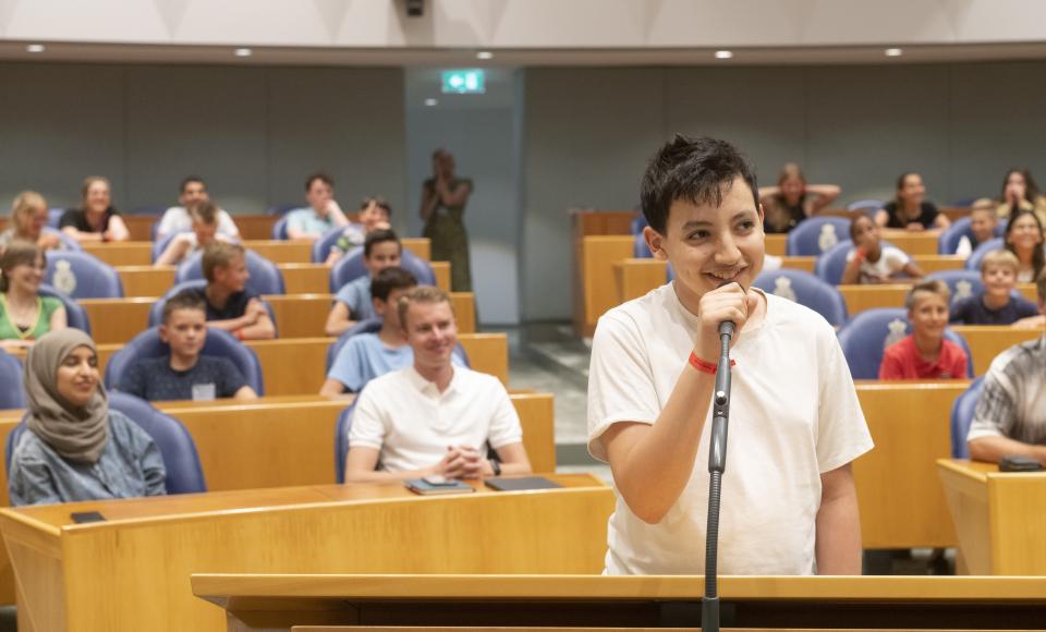 Een jongere staat in de plenaire zaal bij de interruptiemicrofoon. In de bankjes achter hem zitten twee Kamerleden en jongeren.