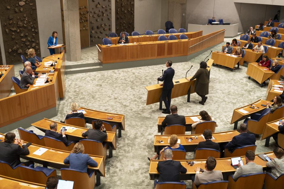 Plenaire zaal van bovenaf gezien met links de Voorzittersstoel in het midden twee Kamerleden aan de interruptiemicrofoon, aan de onderkant Kamerleden op de rug gezien en bovenin de foto in de verte het zogeheten vak K, waar de bewindspersonen zitten. 