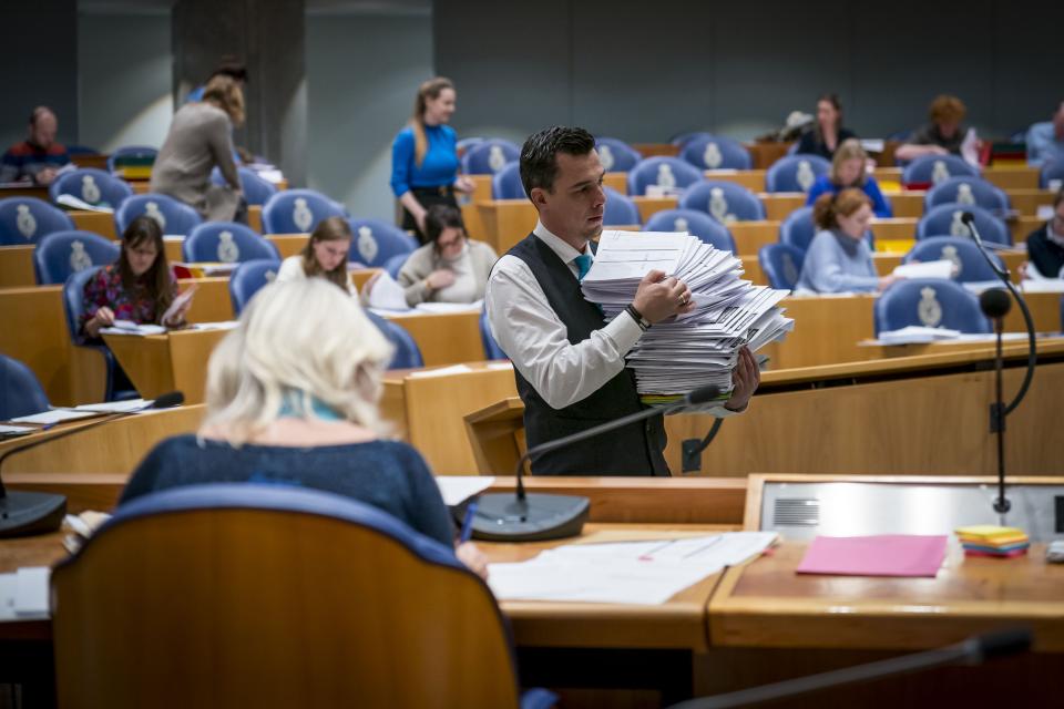 Mensen in de plenaire zaal nemen de Kamerbankjes in beslag en lopen rond met documenten. 