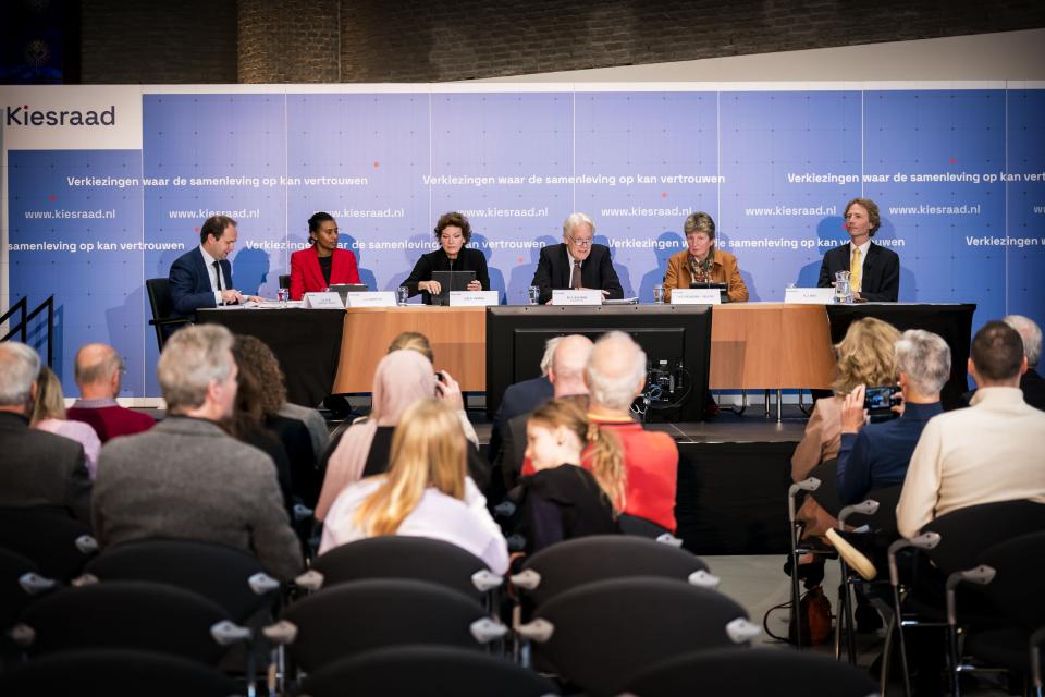 Zes mensen achter een lange tafel op een podium met het gezicht naar de zaal. Op de rug gezien mensen in stoelen die richting het podium kijken. Achterwand met logo's van de Kiesraad. 