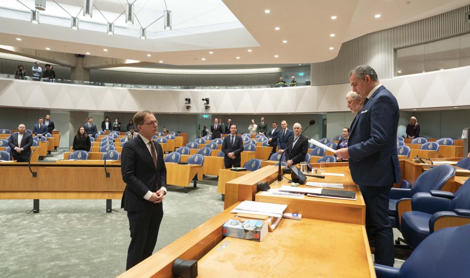 Pepijn van Houwelingen staat voor het bureau van Kamervoorzitter Bosma die met een blaadje in zijn hand neerkijkt op het Kamerlid. Op het bureau liggen boeken. In de achtergrond staan verscheidene Kamerleden voor hun zetel.