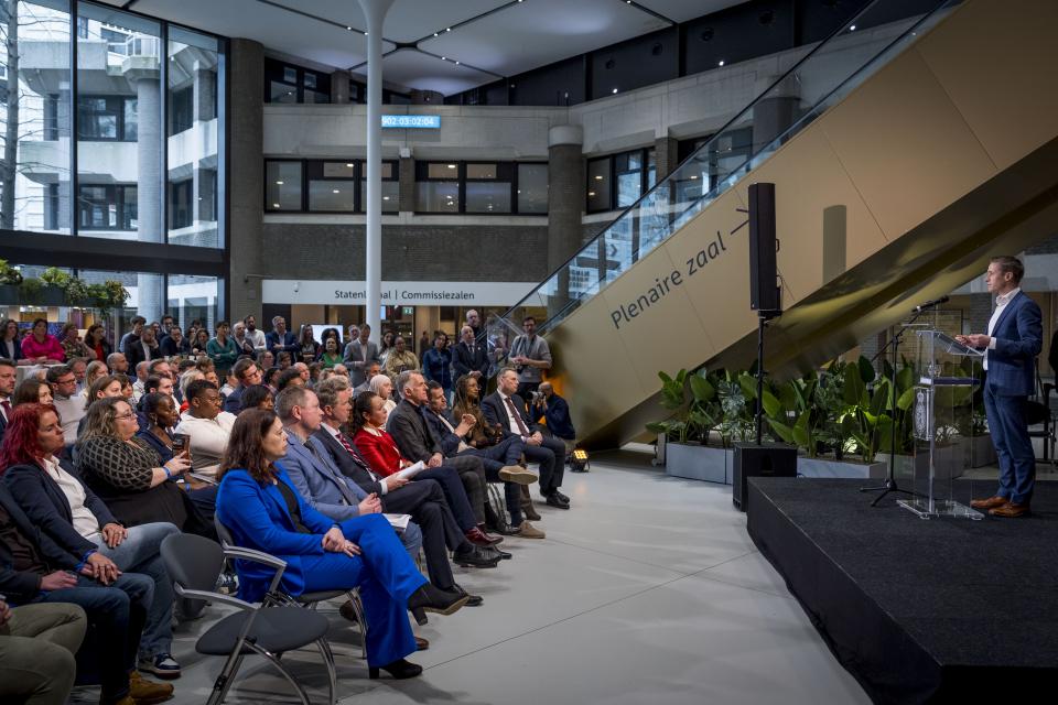 In de Statenpassage van de Tweede Kamer, met op de achtergrond de roltrap, kijkt publiek op stoeltjes naar commissievoorzitter Michiel van Nispen, die een toespraak houdt bij de presentatie van het rapport.