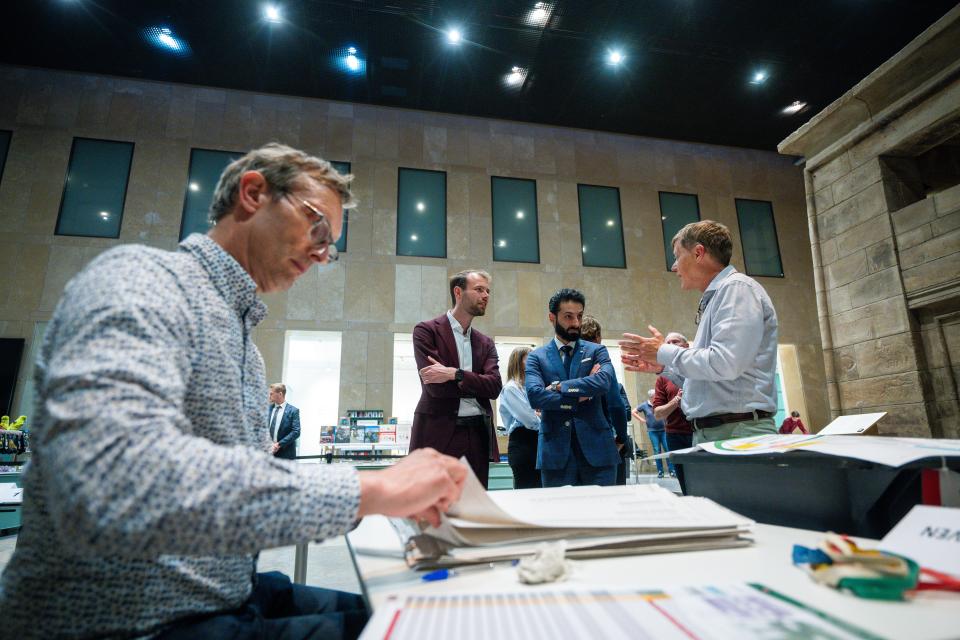 Op de voorgrond een man achter een tafel die formulieren voor zich heeft, daarachter staan twee mannen die uitleg krijgen van een andere man. 