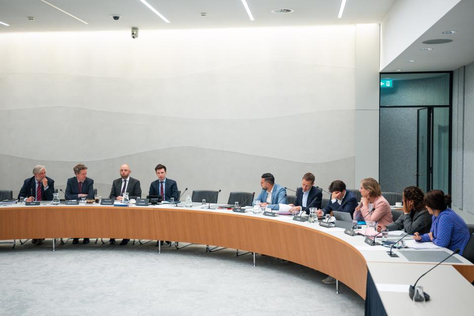 Personen zitten aan een lange tafel. Op de tafel staan microfoons en liggen documenten. 