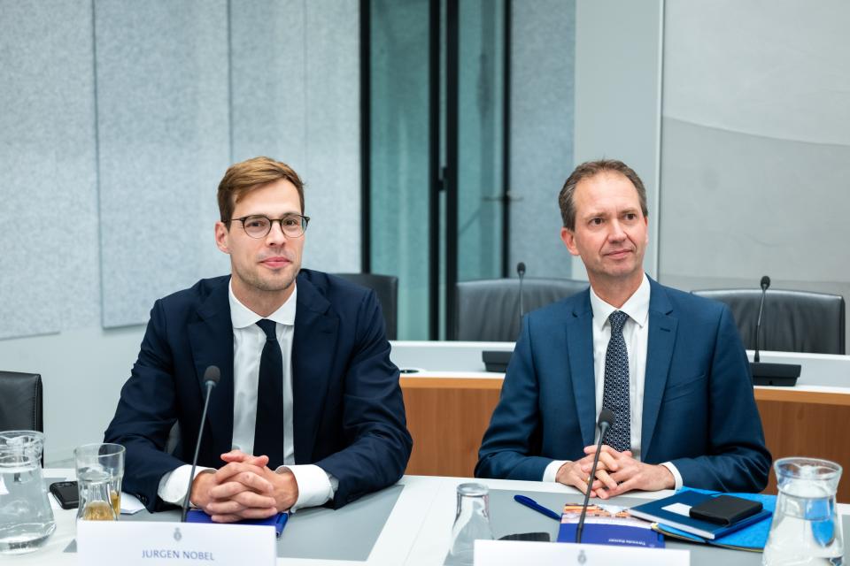 Twee mannen (Jurgen Nobel en Eddy van Hijum) zitten naast elkaar aan tafel, allebei met de handen gevouwen, ieder achter een microfoon. 