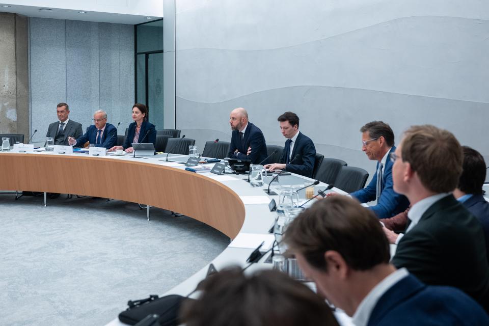 Achter een lange tafel in halve cirkel opstelling zitten links de drie kandidaat-bewindspersonen, daarnaast de commissievoorzitter en de -griffier, rechts in beeld en het dichtstbij de commissieleden van opzij gezien. 