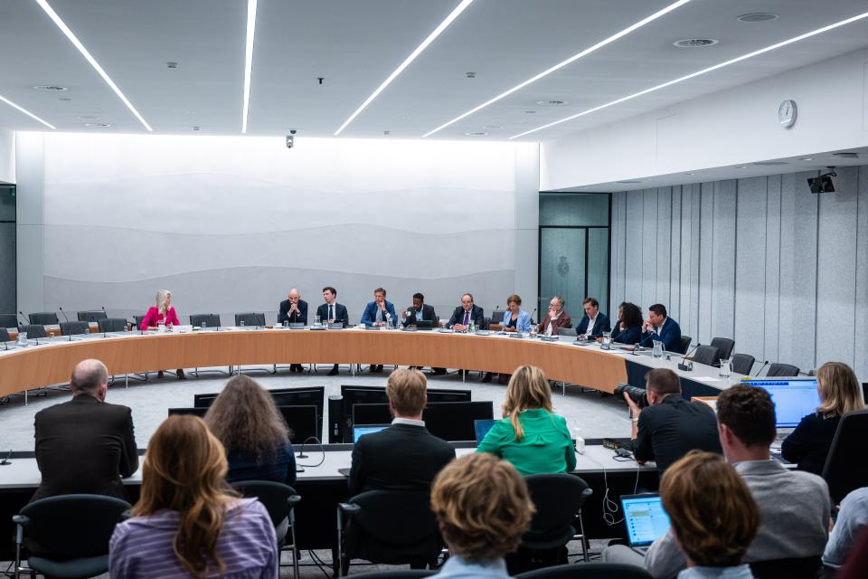 De vergaderzaal met tegen de achterwand de kandidaat-minister en de commissieleden. Op de voorgrond, op de rug gefotografeerd, het publiek op de perstribune.