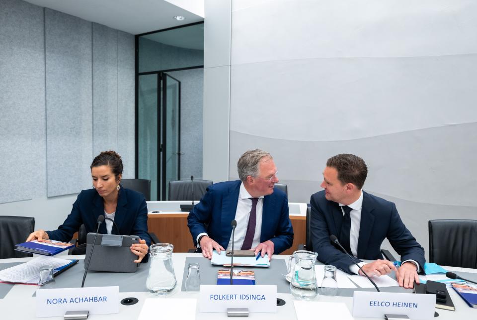 De drie kandidaat-bewindspersonen van Financiën zitten naast elkaar aan een tafel achter microfoons. Twee kandidaten praten met elkaar.  