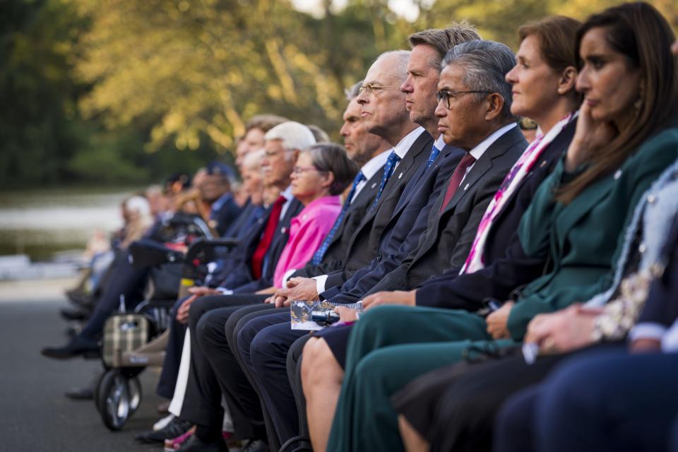 De eerste rij van genodigden op stoelen tegen een achtergrond van bomen en water. Zij luisteren naar een van de sprekers bij het Indisch Monument. 