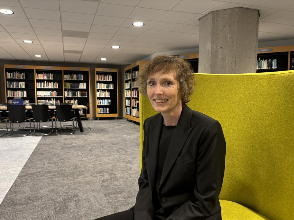 Kenniscoördinator Susan van Velzen zit op een gele bank met daarachter boekenkasten.