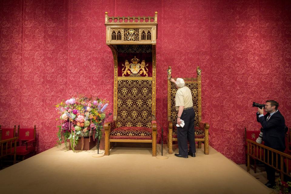 Op het podium in de Koninklijke Schouwburg staat links een groot boeket bloemen, daarnaast de troon en de zetel voor de Koningin. Daarvoor is een man, op de rug gezien, met een doek bezig met afstoffen.  