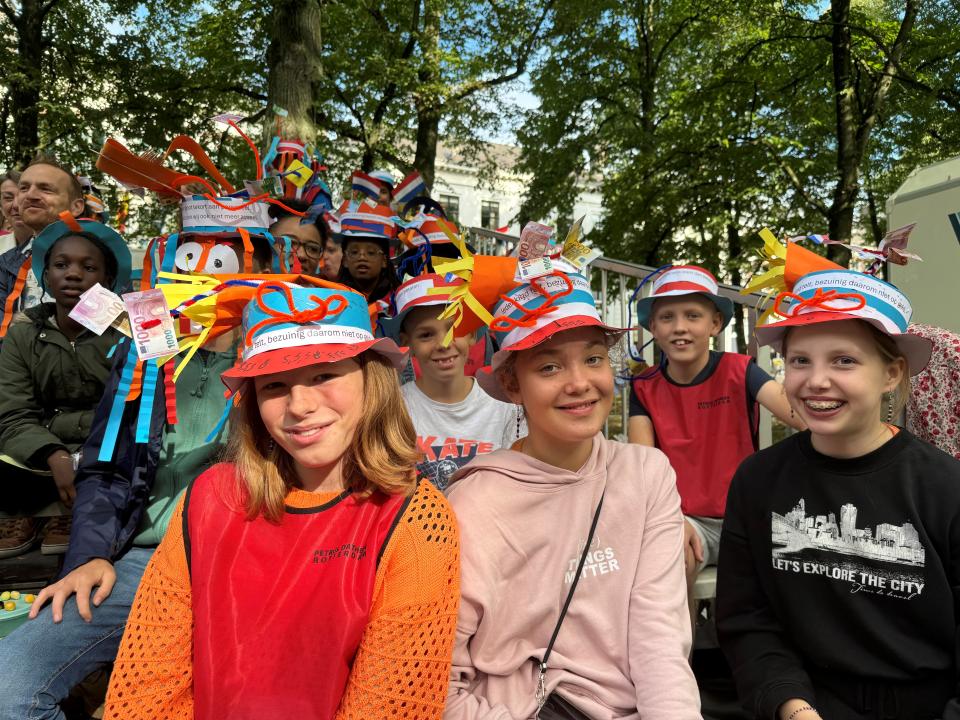 Een groep kinderen met kleurrijke kleding en hoedjes op de publiekstribune bij de Koninklijke Schouwburg.  