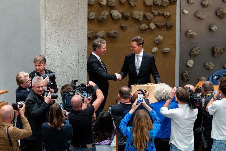 Kamervoorzitter Martin Bosma schudt de hand van minister Eelco Heinen van Financiën in de plenaire zaal, in het vak waar de bewindspersonen zitten. Op de voorgrond fotografen op de rug gezien, die de twee op beeld zetten.  