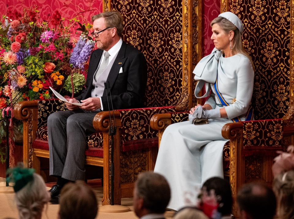 De Koning leest de troonrede voor. Achter hem en de Koningin een rode wand, voor hen de Kamerleden, kabinetsleden en gasten van achteren gefotografeerd. 