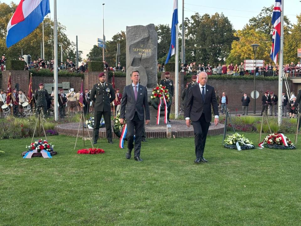 De twee Kamervoorzitters lopen over een grasveld terug naar hun plek. Achter hen de krans die zij zojuist hebben gelegd, een rij vlaggen en het gedenkteken in steen. 