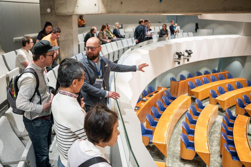 Een man wijst een groepje bezoekers vanaf de publieke tribune onderdelen van de plenaire zaal aan. 