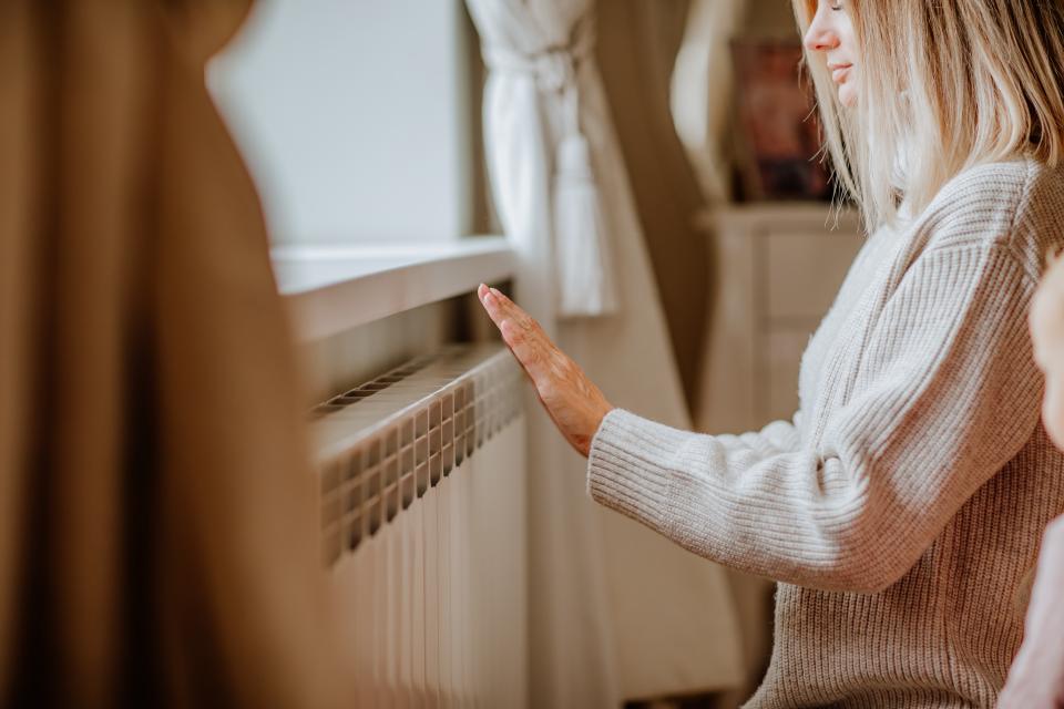 Blonde vrouw in beige trui warmt haar handen aan een radiator.