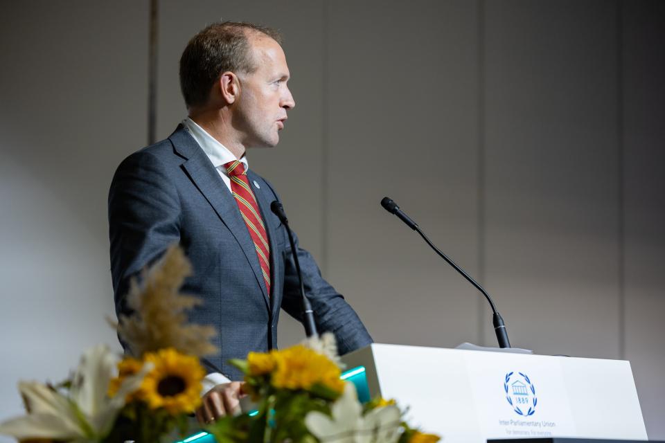 Op de foto staat Eerste Kamerlid en delegatieleider de heer Hendrik-Jan Talsma. Hij spreekt vanachter ene katheder en op de voorgrond is een boeket bloemen zichtbaar.