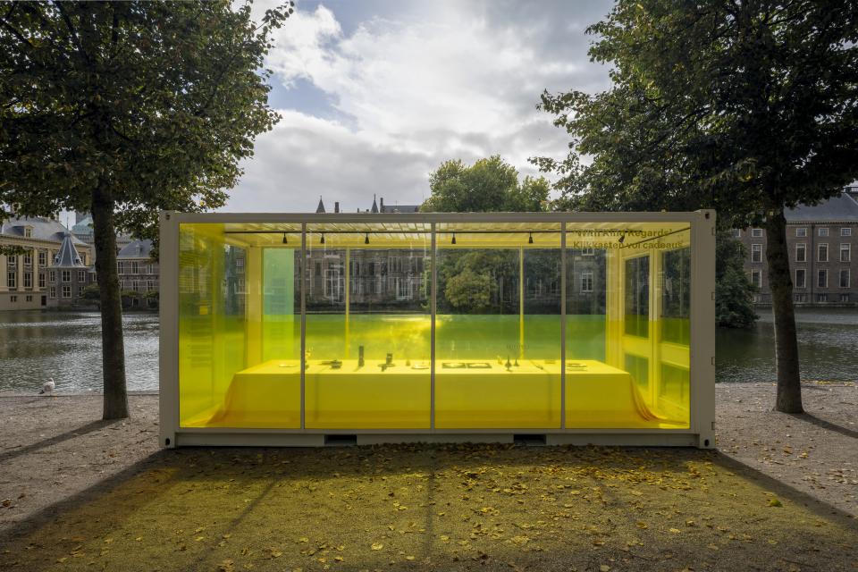 Geel verlichte glazen container staat tussen de bomen langs de Hofvijver. 