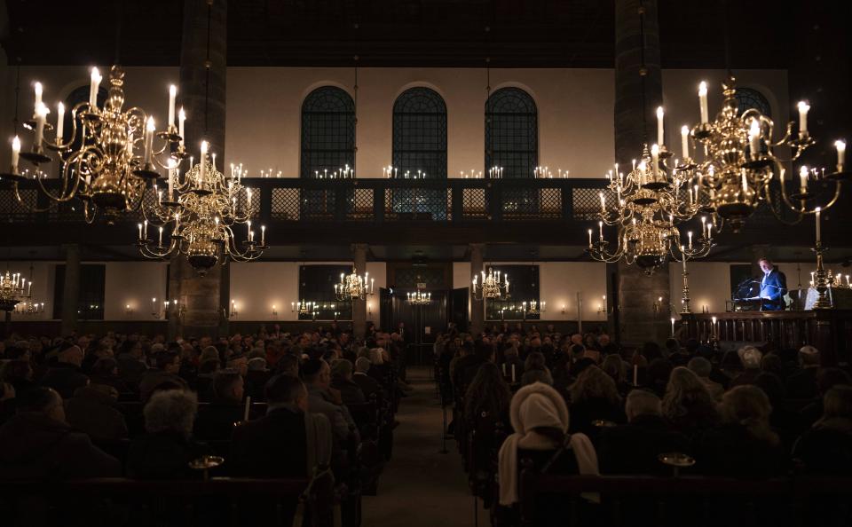 Interieur Portugese Synagoge. Links en rechts grote kroonluchters, daartussen in halfduister publiek op de rug gezien. Rechts in de verte is een spreker uitgelicht. 