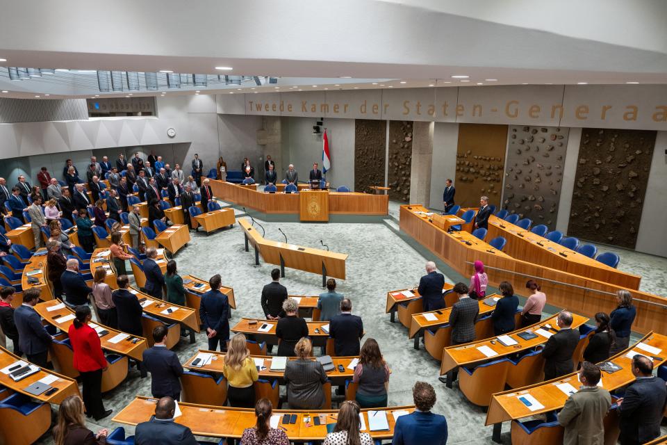 Kamerleden en andere aanwezigen staan in de plenaire zaal tijdens de herdenking van Hans van den Broek. Kamervoorzitter Martin Bosma leest zijn herdenkingsspeech voor. In vak K staan minister-president Dick Schoof en minister van Sociale Zaken en Werkgelegenheid Eddy van Hijum.