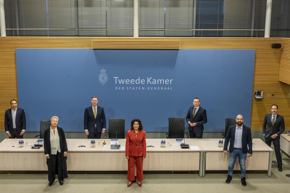 Kameleden staan afwisselend voor en achter een tafel in de Enquêtezaal van de Tweede Kamer. Van links naar rechts zijn het Maarten Groothuizen (D66), Corrie van Brenk (50PLUS), André Bosman (VVD, voorzitter), Nevin Özütok (GL, ondervoorzitter), Evert Jan Slootweg (CDA), Cem Laçin (SP) en John Kerstens (PvdA).