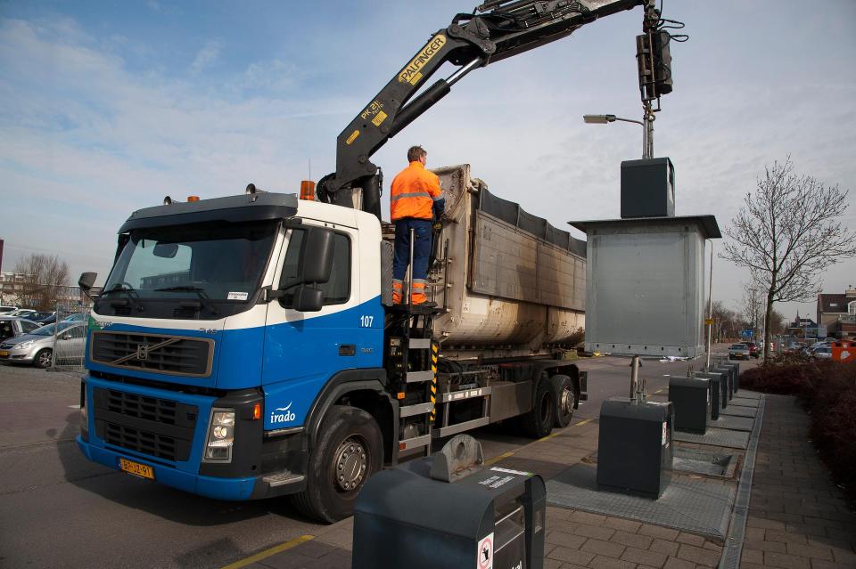 Een vrachtwagen met grijparm rechts trekt een straatcontainer omhoog. 