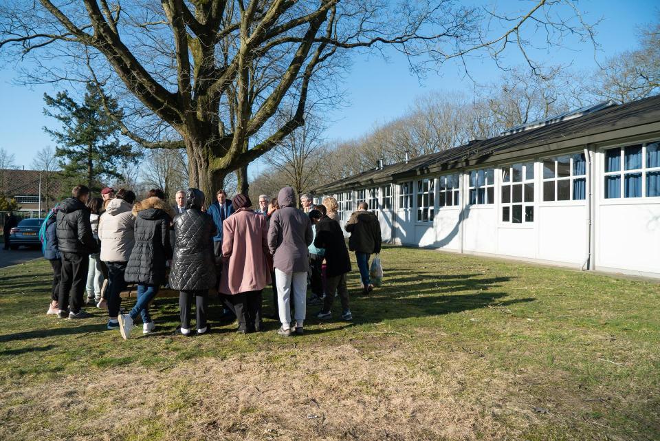 Een groep mensen op de rug gezien, achter hen een boom en rechts een rij witte houten woningen.