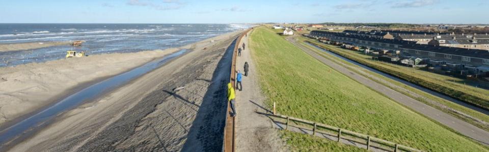 Dijk van bovenaf gefotografeerd: links eerst strand en dan water, rechts eerst gras en dan huizen. 