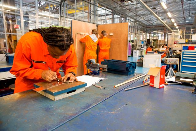 Jongeren in oranje overalls. Vooraan buigt een van hen zich over een werktafel, daarachter staan twee studenten met de rug naar de camera bij een houten scheidingswand.