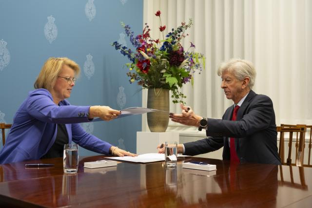 Kamervoorzitter Roelien Kamminga (links) en verkenner Ronald Plasterk zitten ieder aan een kant van een tafel en wisselen documenten uit. 