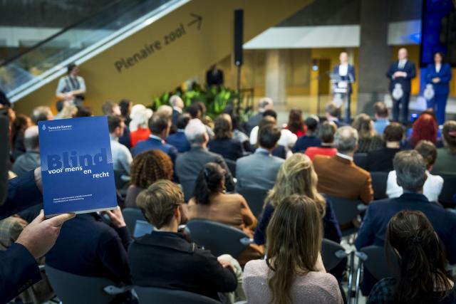 Op de voorgrond houdt een hand het rapport Blind voor mens en recht vast. Op de achtergrond de Statenpassage met toeschouwers die zitten op stoelen en een podium waarop drie commissieleden staan.