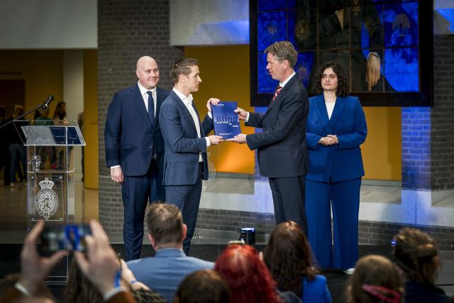 Op een podium de drie leden van de enquêtecommissie plus in het midden Kamervoorzitter Bosma, die van Michiel van Nispen het rapport overhandigd krijgt. 