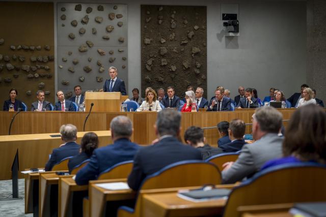 Minister-president Dick Schoof staat achter het spreekgestoelte in het zogeheten vak K, waar ook alle andere bewindspersonen aanwezig zijn. Op de voorgrond Kamerleden op de rug gezien. 