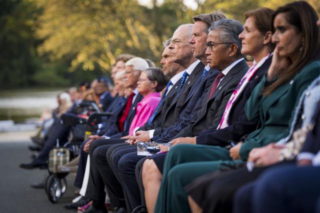 De genodigden naast elkaar zittend op de eerste rij tegen een achtergrond van bomen en water, luisteren naar een van de sprekers bij het Indisch Monument. 