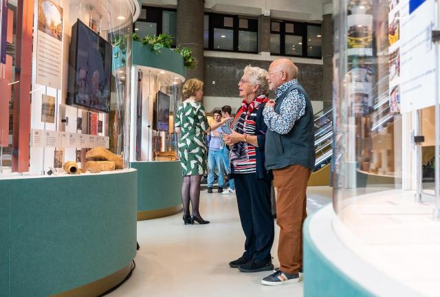 Twee mensen, van de zijkant gezien, kijken in een glazen vitrine die onderdeel uitmaakt van de tentoonstellingen in de Tweede Kamer. 